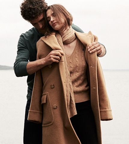 Man draping camel coat over woman's shoulders on beach